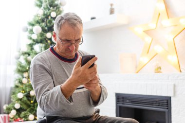 Noel, tatiller ve insanlar konsepti. Mutlu gülümseyen yaşlı adam evde akıllı telefonuyla oturuyor.