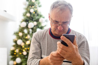 Noel, tatiller ve insanlar konsepti. Mutlu gülümseyen yaşlı adam evde akıllı telefonuyla oturuyor.