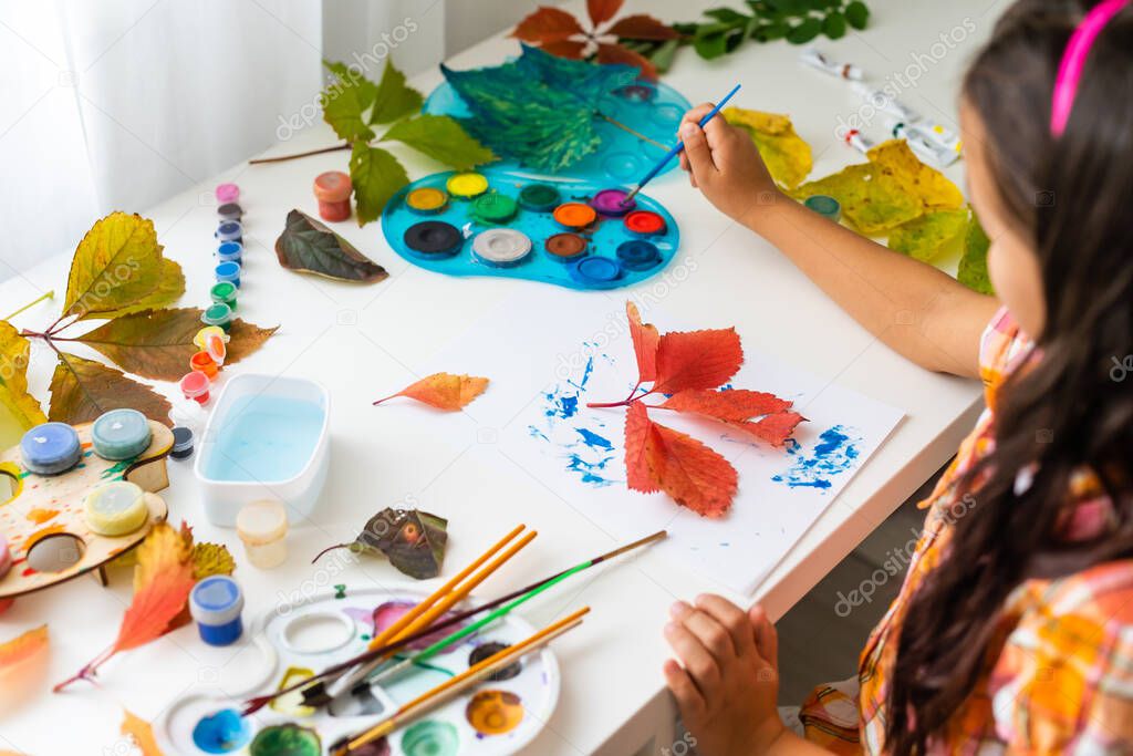 little girl paints leaves with brush at home, autumn art.