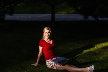 Parkta dizüstü bilgisayar kullanarak gülümseyen genç kadın.