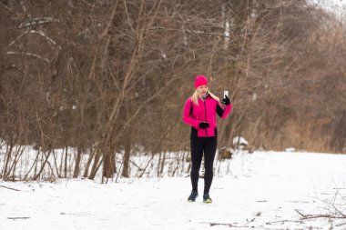 Siyah eşofmanlı kızıl kız koşuyor ve telefonuna bakıyor. Arka planda kış manzarası. - Evet. Yüksek kalite fotoğraf