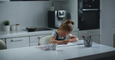 4-7 yaşlarında bir kız çocuğu evde oturuyor ve kağıda resim yapıyor. Çocuk resme odaklanmış bir gökkuşağı çiziyor. Çocuk çizimin kötü sonuçlanacağı için üzgün. Kadın portresi.