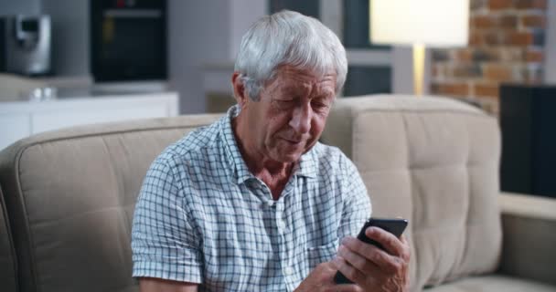 Viejo caucásico sostiene en su mano el teléfono inteligente con expresión de dificultad en su uso. Comunicación con familiares a través de video comunicación . — Vídeos de Stock