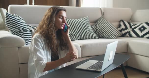 A young brunette woman sits in a room and is working on a laptop, typing text. The mobile phone rang, the woman answered the call and started talking. — Stock Video