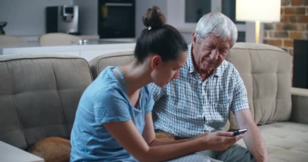 Sorprendido padre barbudo mayor usando teléfono inteligente mientras estaba sentado en el sofá gris con su hija adulta, se sorprendió de las tecnologías modernas. La hija enseña a su padre a usar un teléfono móvil . — Vídeos de Stock