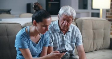 Aydınlık oturma odasındaki gri kanepede yetişkin esmer kızıyla oturan yaşlı ve sakallı babayı şaşırttı ve modern teknolojileri şok etti. Kız babasına öğretiyor. 