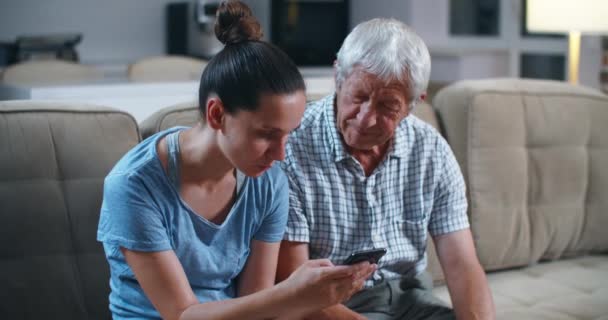 Sorprendido padre barbudo mayor mientras estaba sentado en el sofá gris en la sala de estar con su hija adulta, se sorprendió de las tecnologías modernas. La hija le enseña a su padre a usar el teléfono . — Vídeos de Stock