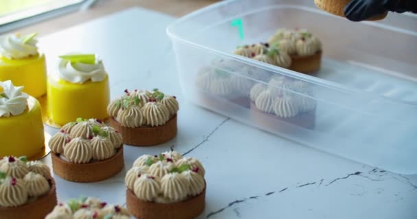 En la mesa hay dulces postres frescos preparados para la venta. Los postres se transfieren a un contenedor para su transporte. Primer plano de las manos femeninas en guantes negros y postres — Vídeos de Stock