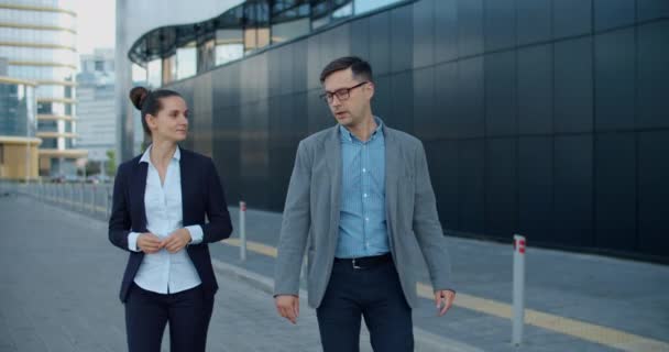 Twee collega 's kletsen op straat tijdens de lunchpauze in de buurt van het gebouw. Ze lopen langs een moderne straat vol met glazen gebouwen en structuren. Communicatie van collega 's — Stockvideo