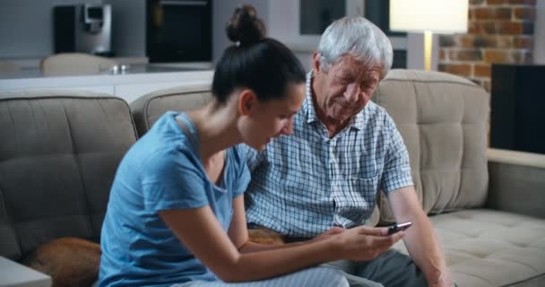 Sorprendido padre barbudo mayor mientras estaba sentado en el sofá gris en la sala de estar con su hija morena adulta, se sorprendió de las tecnologías modernas. La hija enseña a su padre — Vídeos de Stock