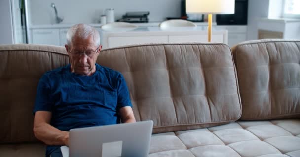 Ein älterer Mann von 60 Jahren mit Brille sitzt auf dem Sofa und schreibt auf einem Laptop Nachrichten an seine Kinder. Porträt eines älteren Mannes. — Stockvideo