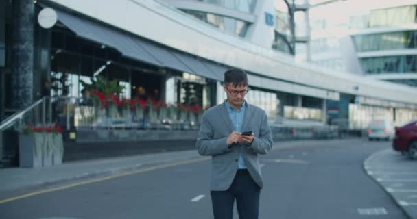 Jonge mannelijke zakenman loopt moderne straat en sms 't collega' s. Een belangrijke onderhandeling. Tegen de achtergrond van een moderne straat met glazen gebouwen en wegen. — Stockvideo