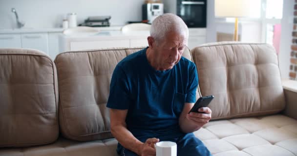 Boodschap van kinderen. Een vrolijke grijze man die zijn telefoon vasthoudt met het lezen van een boodschap van kinderen en thee drinken. Vreugde voor een oudere persoon. — Stockvideo