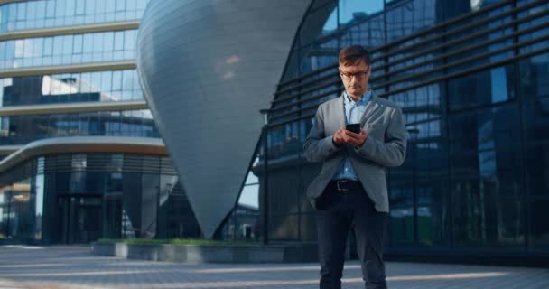 Oung mannelijke zakenman staat op een moderne straat en praat op een mobiele telefoon met collega 's. Een belangrijk telefoontje. Tegen de achtergrond van een moderne straat met glazen gebouwen. — Stockvideo