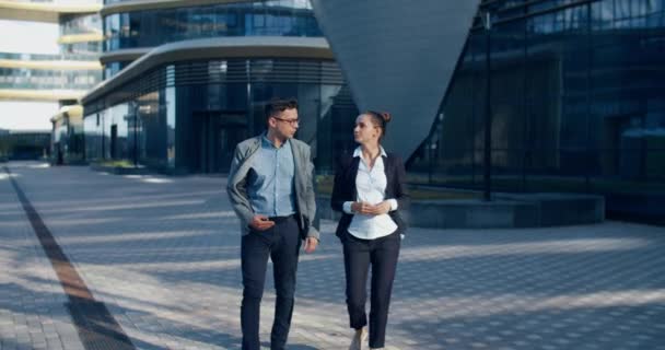 Twee collega 's kletsen op straat tijdens de lunchpauze in de buurt van het gebouw op de achtergrond. Ze lopen langs een moderne straat vol met glazen gebouwen en structuren. Communicatie van collega 's — Stockvideo