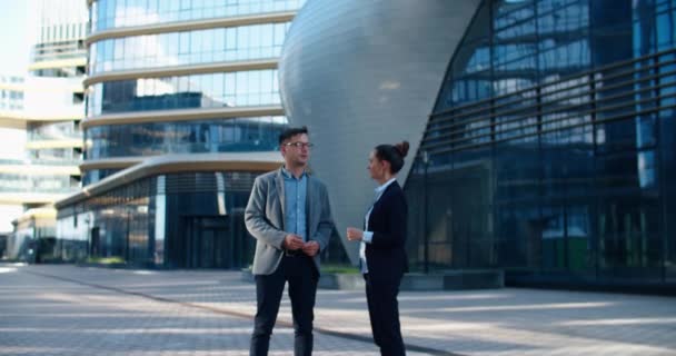 Twee collega 's kletsen op straat tijdens de lunchpauze in de buurt van het gebouw op de achtergrond. Ze staan in een moderne straat vol met glazen gebouwen en structuren. Communicatie van collega 's — Stockvideo