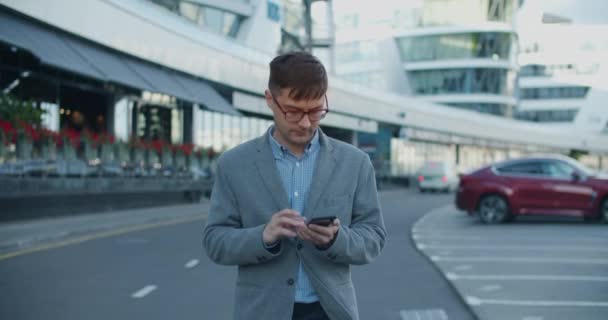 Young male businessman walks modern street and writes texting colleagues on mobile. An important negotiation. Against the background of a modern street with glass buildings and roads. — Stock Video