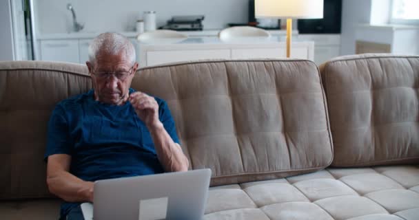 Un anciano de 60 años con gafas se sienta en el sofá y escribe mensajes a sus hijos en un portátil, está cansado, se quita las gafas y hace un masaje facial. — Vídeos de Stock