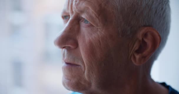 Un retrato de cerca de un anciano de unos 60 años que se ve triste por la ventana. Pensamientos sobre la vejez — Vídeos de Stock