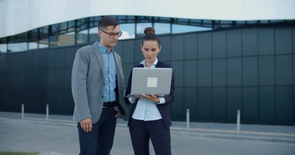 Dwóch menedżerów, mężczyzna i kobieta, stoją na ulicy na tle nowoczesnego budynku i omawiają biznes plan, zaglądają do laptopa. Plany pracy na tydzień. Dyskusje robocze. — Wideo stockowe