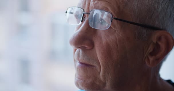 Un retrato de cerca de un anciano con gafas, que se ve triste por la ventana. Se quita las gafas. Pensamientos sobre la vejez — Vídeos de Stock