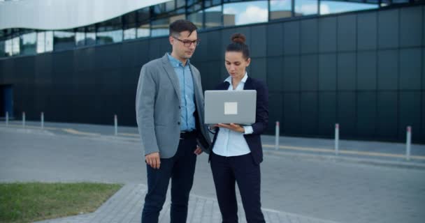 Zwei Manager, ein Mann und eine Frau, stehen auf der Straße vor dem Hintergrund eines modernen Gebäudes, diskutieren einen Businessplan, blicken in einen Laptop. Arbeitspläne für die Woche. Arbeitsgespräche. — Stockvideo
