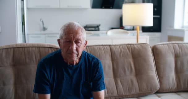 Een oudere man met droevige ogen zit op de bank in de kamer en reflecteert op het leven en de ouderdom. Op de achtergrond een keuken en een lamp. Mannelijk portret — Stockvideo