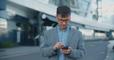 İş stili olan modern bir adam cam bir binanın arka planında dikiliyor ve telefonda bir mesaj yazıyor. Gözlük takan işadamı iş sorunlarını telefonla çözüyor