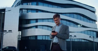 İş stili olan modern bir adam cam bir binanın arka planında dikiliyor ve telefonda bir mesaj yazıyor. Gözlük takan işadamı iş sorunlarını telefonla çözüyor