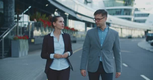 Dwóch menedżerów, mężczyzna i kobieta, spacerują po ulicy na tle nowoczesnego budynku i omawiają biznes plan podczas spaceru po mieście. Plany pracy na tydzień. Dyskusje robocze. — Wideo stockowe