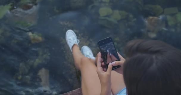 Ευτυχισμένη γυναίκα κάθεται σε μια προβλήτα στο βάθος της θάλασσας και φωτογραφίζει τα πόδια της στο τηλέφωνο. Αναψυχή και τουριστική έννοια. Άποψη από ψηλά — Αρχείο Βίντεο