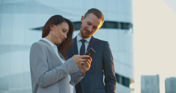 Alegre sonriente joven pareja de colegas de negocios de pie cerca del centro de negocios en la calle utilizando teléfonos móviles. Comunicación de colegas en la calle. Retrato de una mujer y un hombre — Vídeo de stock