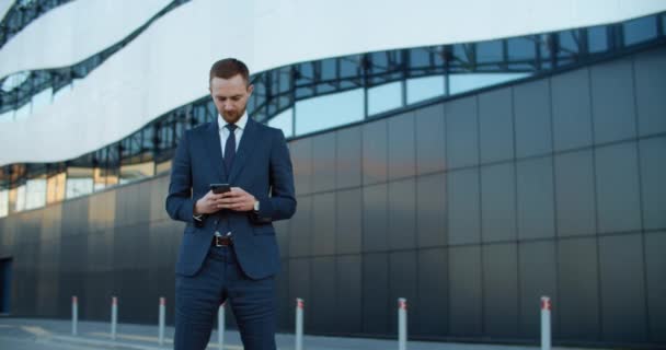 ビジネス用の服を着た若い男が近代的な建物の背景に立って電話でメッセージを書きます。仕事の対応. — ストック動画