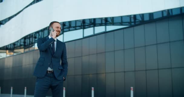 Een jonge man in zakelijke kleding staat tegen de achtergrond van een modern gebouw en praat aan de telefoon. Werkoproep. — Stockvideo