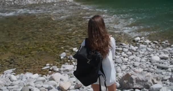 Sullo sfondo di un fiume di montagna si staglia una donna sportiva con una camicia bianca e uno zaino. Il fiume scorre sulle pietre. Vista posteriore — Video Stock
