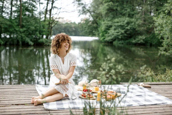 Happy young woman with curly hair sits in the park on a blanket. A woman on a picnic, on a blanket, a lot of different delicious food and drinks. Summer picnic. Womens rest with reading a book.