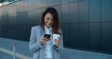 İş stili olan güzel bir kadın sokakta duruyor ve telefona bir mesaj yazıyor, kahve içiyor ve gülüyor. Kadın portresi..