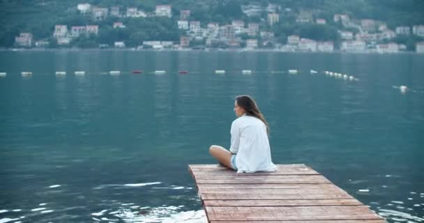 Uma bela turista com cabelo encaracolado senta-se no cais sozinha e reflete sobre diferentes tópicos. Vista da baía e montanhas — Vídeo de Stock