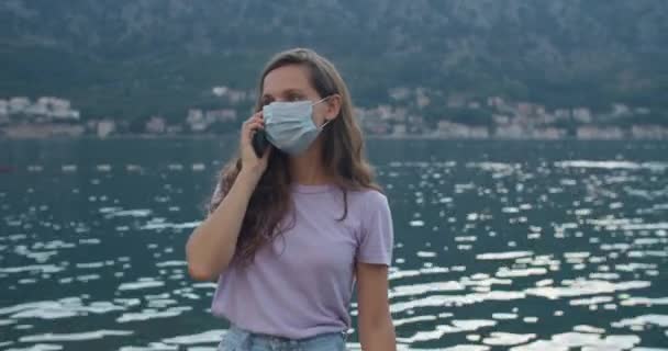 Une jeune femme portant un masque de protection parle au téléphone. Vue sur la baie et les grps. Portrait d'une femme portant un masque de protection. — Video