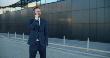 İş adamı takım elbiseli bir adam sokakta duruyor ve modern bir binanın arka planında cep telefonuyla konuşuyor..