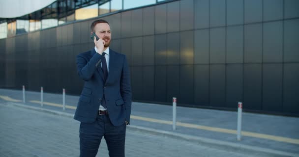 ビジネススーツの男ビジネスマンが通りに立って、現代的な建物を背景に携帯電話で話します. — ストック動画