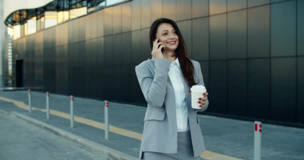 Młoda kobieta w ubraniu biznesowym chodzi na tle nowoczesnego budynku, pije kawę i rozmawia przez telefon. Telefon służbowy. — Wideo stockowe