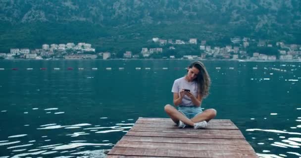 Jovem mulher feliz senta-se em um cais de madeira e escreve uma mensagem em um telefone celular. Vista da baía e das montanhas. Pôr do sol — Vídeo de Stock