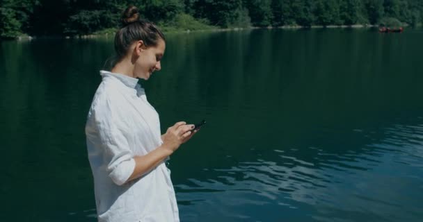 Mulher feliz turista fica em um cais com vista para o lago e montanhas, e escreve uma mensagem ao telefone. Close-up retrato de uma mulher — Vídeo de Stock