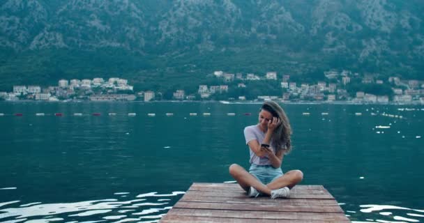 Young happy woman sits on a wooden pier and writes a message on a mobile phone. View of the bay and mountains. Sunset — Stock Video