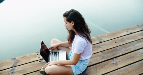Een jonge vrouw zit op de pier en werkt aan een laptop. Freelance. Uitzicht op zee. Het concept van favoriet werk — Stockvideo