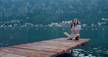 Genç mutlu kadın ahşap bir iskelede oturuyor ve telefonda konuşuyor, gülüyor. Koyun ve dağların manzarası. Gün batımı