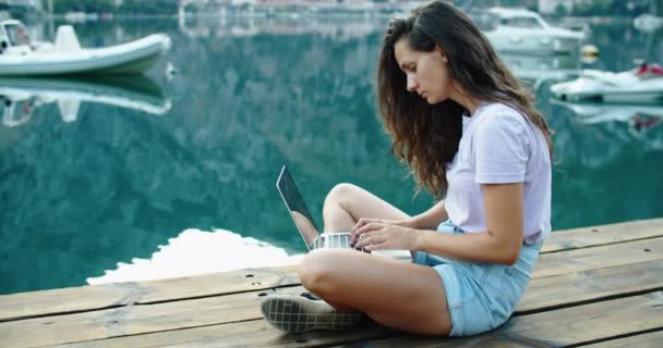 Een jonge vrouw zit op de pier en werkt aan een laptop. Freelance. Uitzicht op zee. Het concept van favoriet werk — Stockvideo