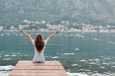 Mutlu kadın turist ahşap bir iskelede oturur ve dinlenir, güzel doğanın tadını çıkarır. Ellerinizi kaldırın. Körfez manzarası ve yeşil dağlar. Arka plan