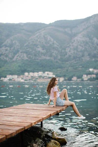 Mutlu kadın turist ahşap bir iskelede oturur ve dinlenir, güzel doğanın tadını çıkarır. Körfez manzarası ve yeşil dağlar.